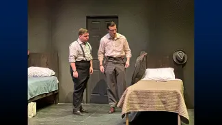 Two actors on stage next to a bed in the performance “The Dumb Waiter” by Harold Pinter.
