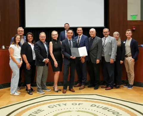 Representatives from 10 colleges across southwestern Wisconsin and northeastern Iowa pose with Greater Dubuque Development Corp. Regional Development Director Nic Hockenberry as he holds a proclamation declaring Aug. 3-7 Community of Colleges week during a city council meeting July 20, 2026.