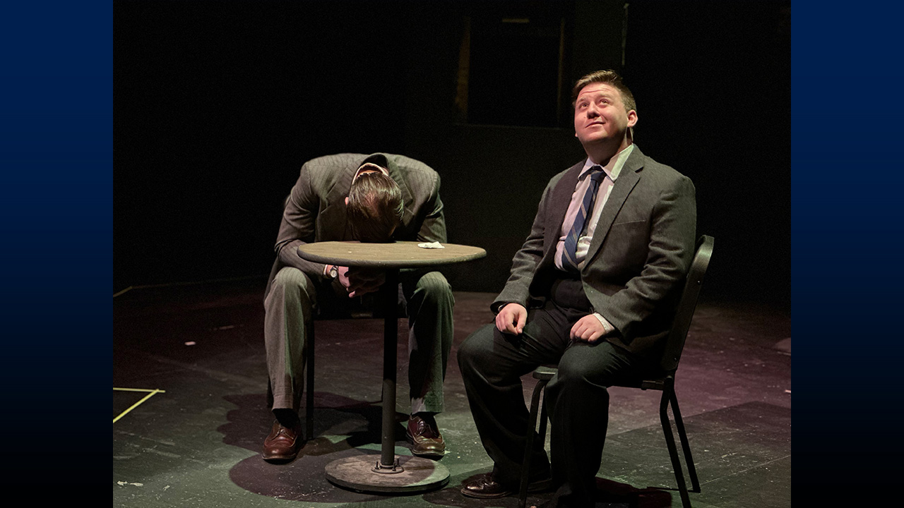 Two actors on stage, sitting at a small round table, one with their head down