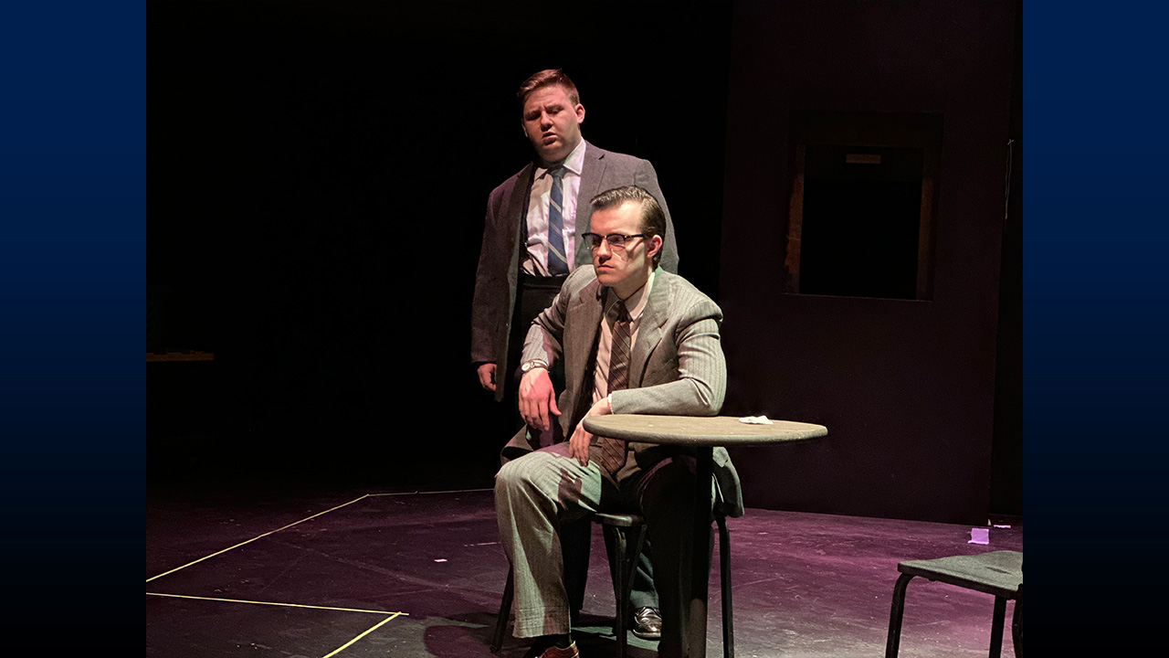 Two actors on stage, one sitting at a small round table and one standing..