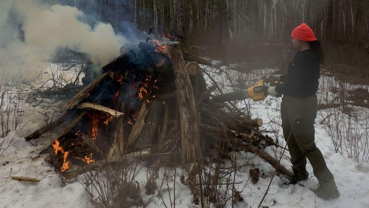 Person wearing a bright orange stocking hat, holding a leafblower and standing near a fire of burning logs