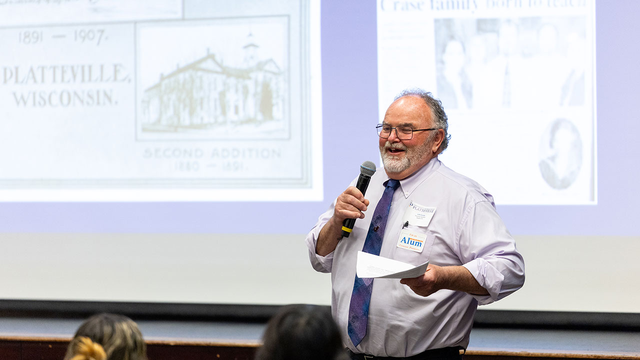Man in a shirt and tie, presenting while smiling and holding a microphone