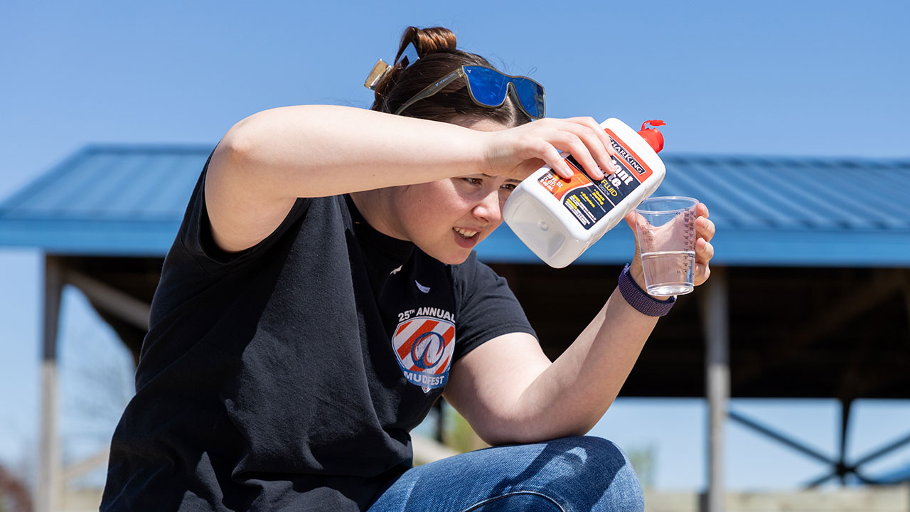 A person outdoors under a clear blue sky, concentrating as they pour liquid from a container into a transparent cup. 