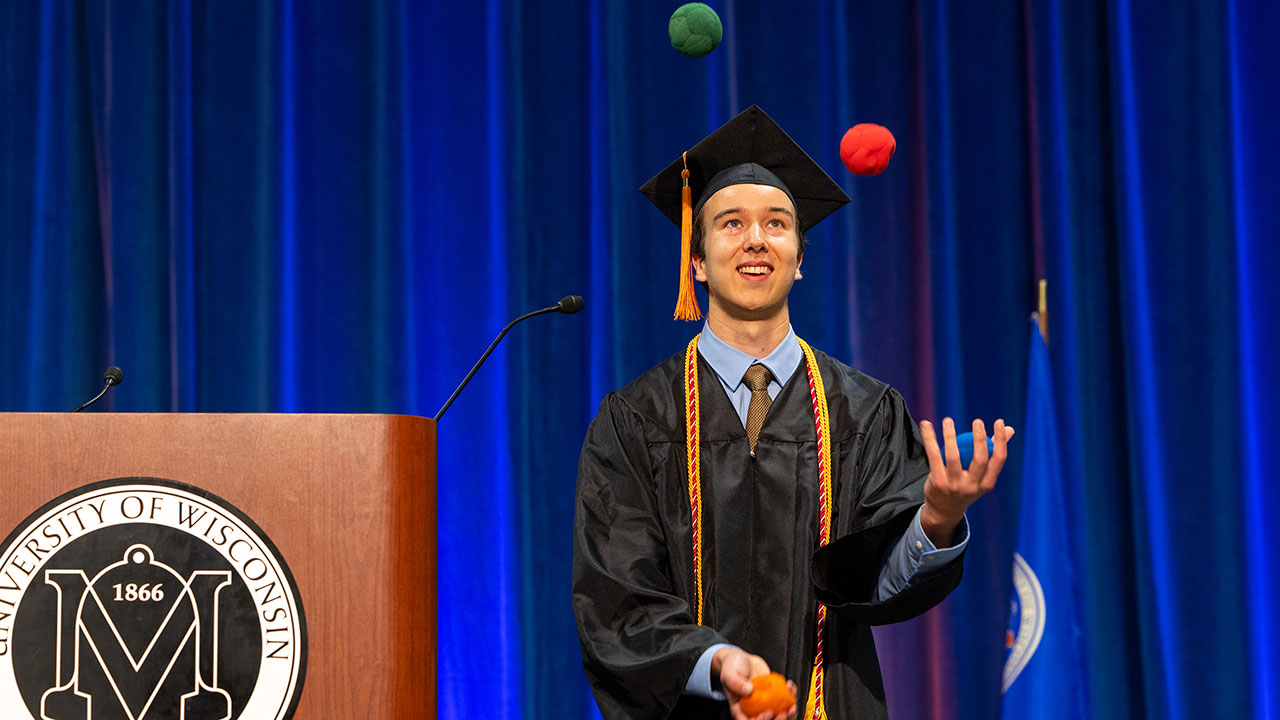 Man in black commencement cap and gown, juggled colorful balls in front of a blue backdrop