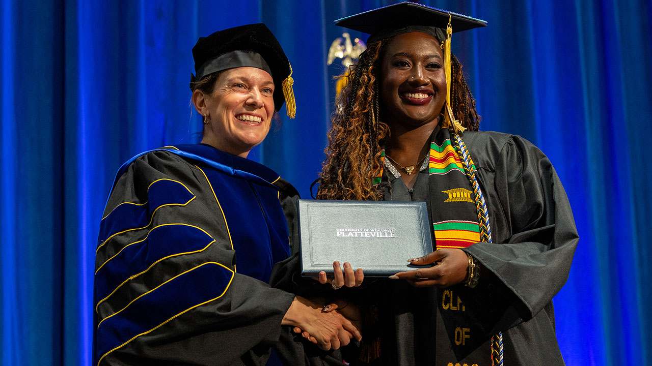 Two women wearing commencement regalia, smiling and holding a diploma