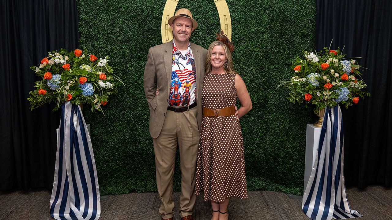 A smiling couple wearing festive clothing pose in front of a Kentucky Derby themed background