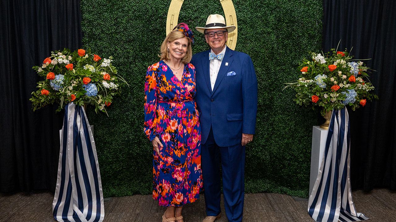 A smiling couple wearing festive clothing pose in front of a Kentucky Derby themed background