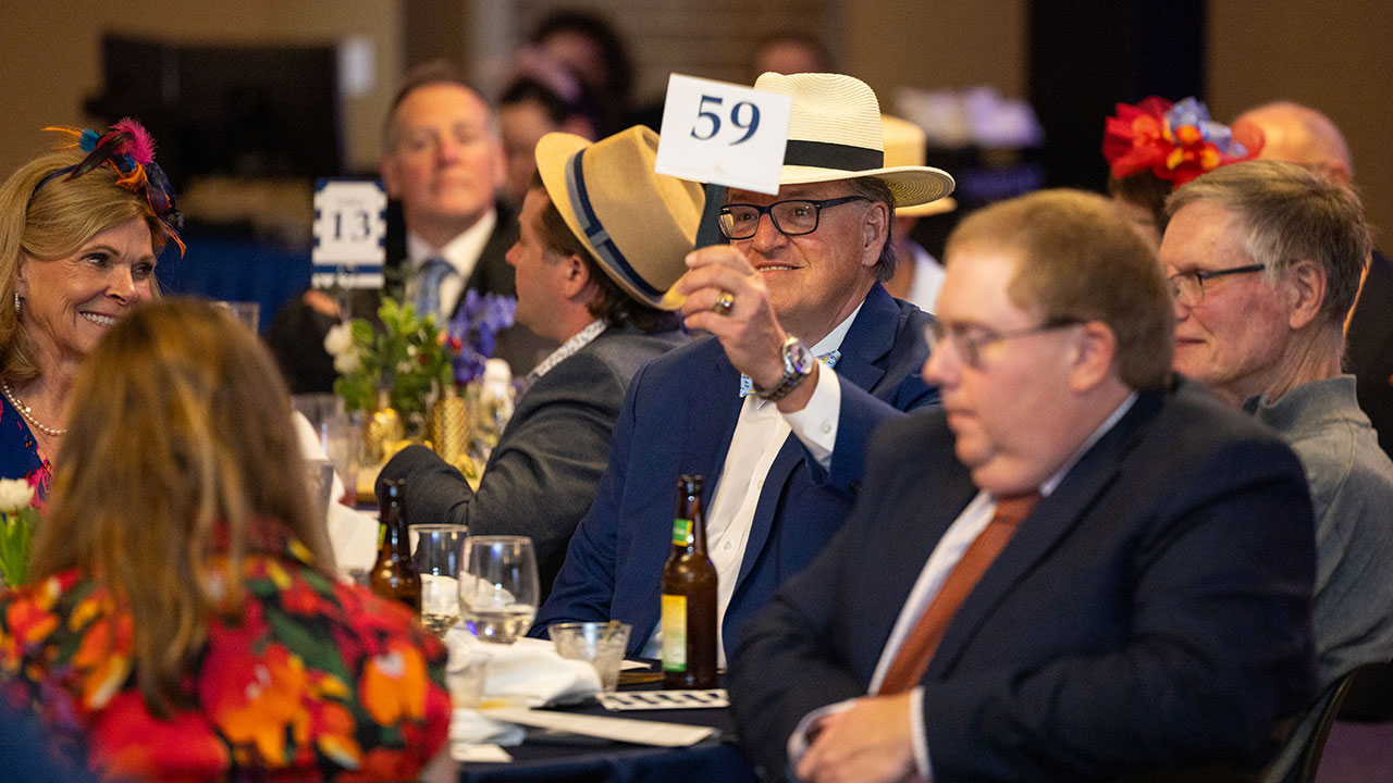 A man seated with others at a round table raises a numbered bidding card during a live auction
