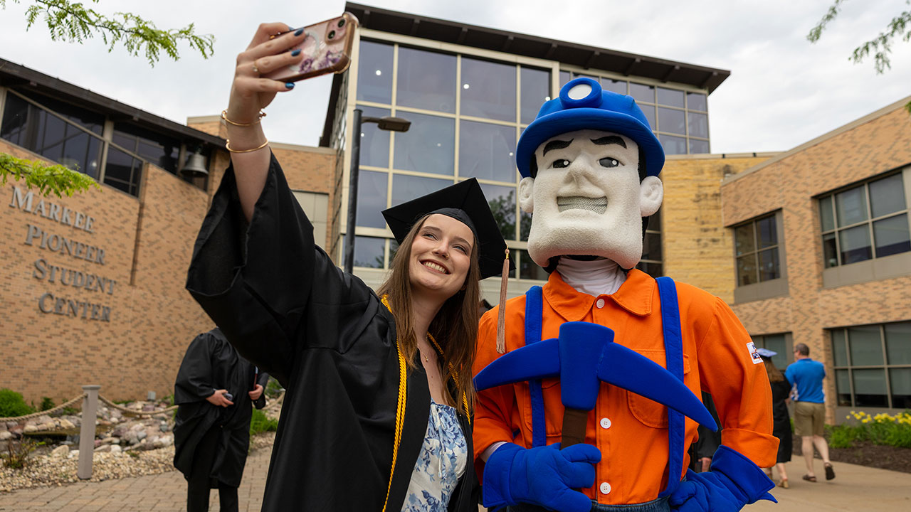 A female student in black cap and gown takes a selfie with a mascot