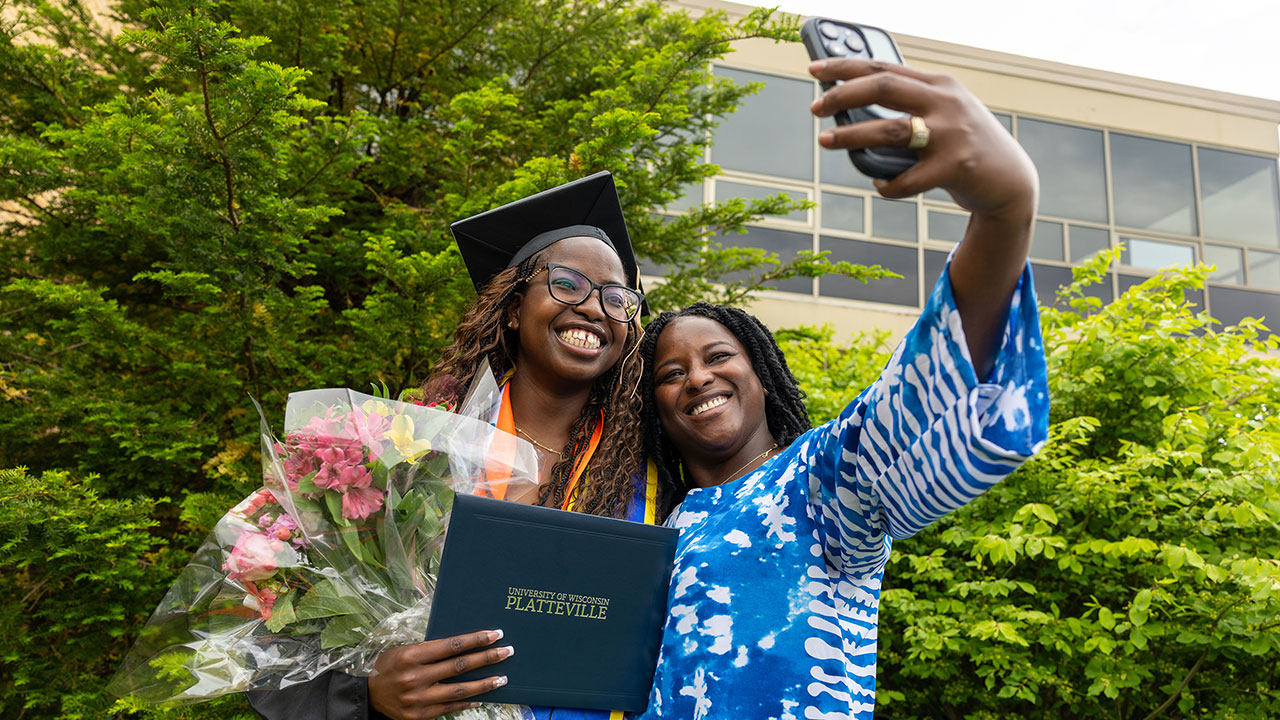 Two females, one wearing a graduation cap and holding a diploma, smiling and taking a selfie