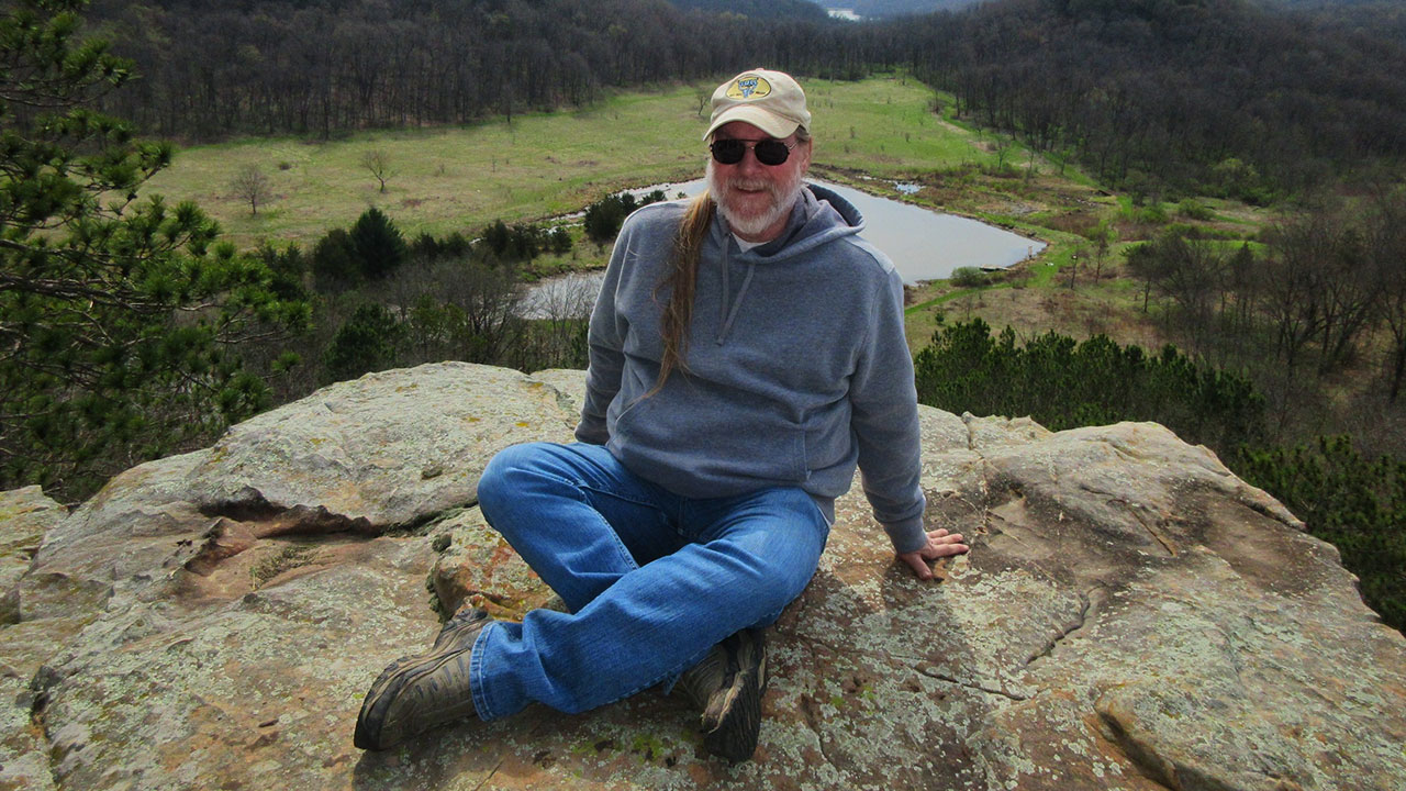 A man wearing a hat sits on a large rock outside