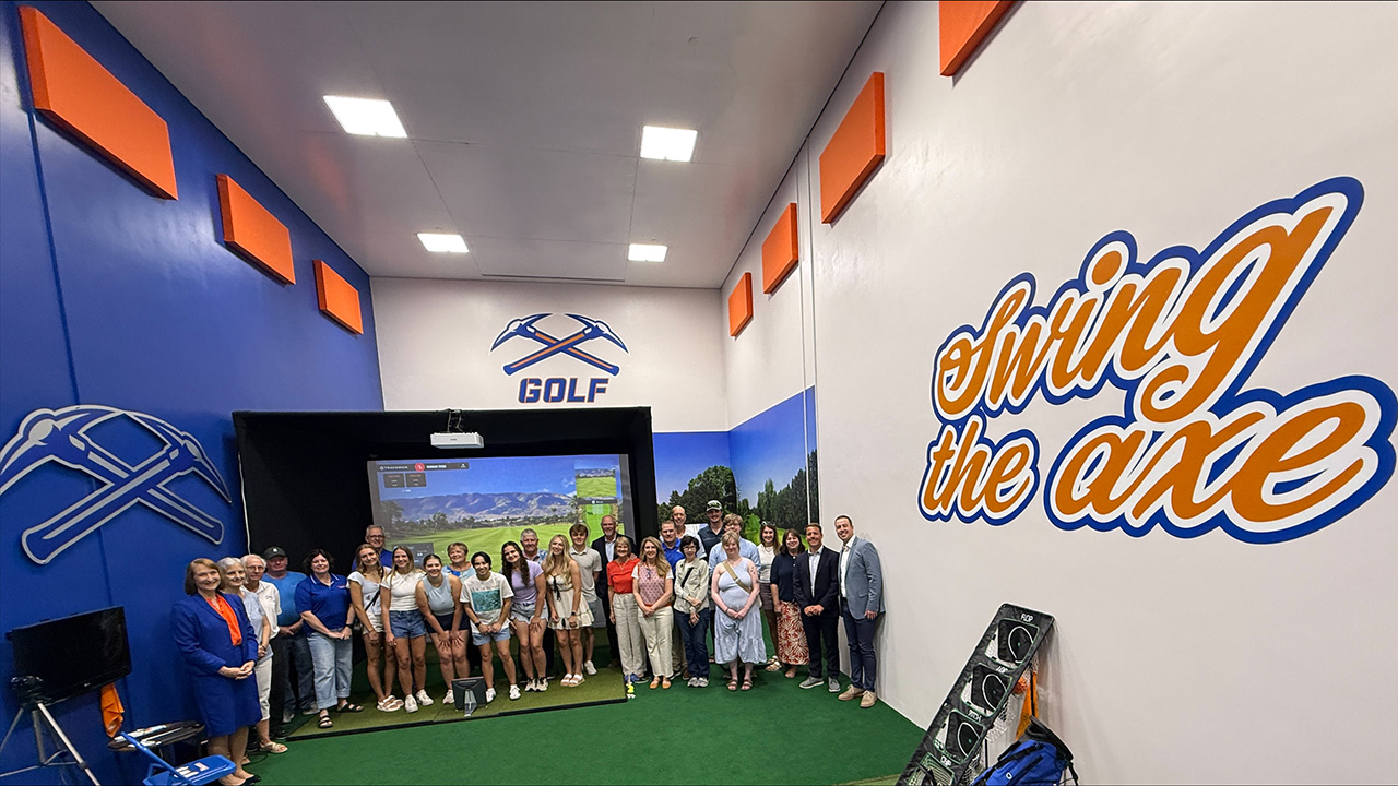 Fish eye view of room with a large group of individuals standing in front of a golf simulator screen.