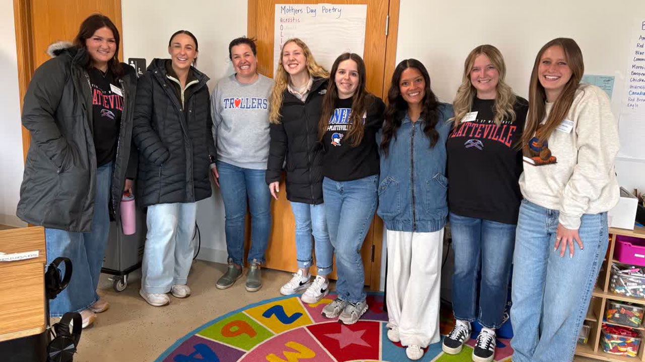 A group of smiling women standing on a colorful rug in an elementary classroom.