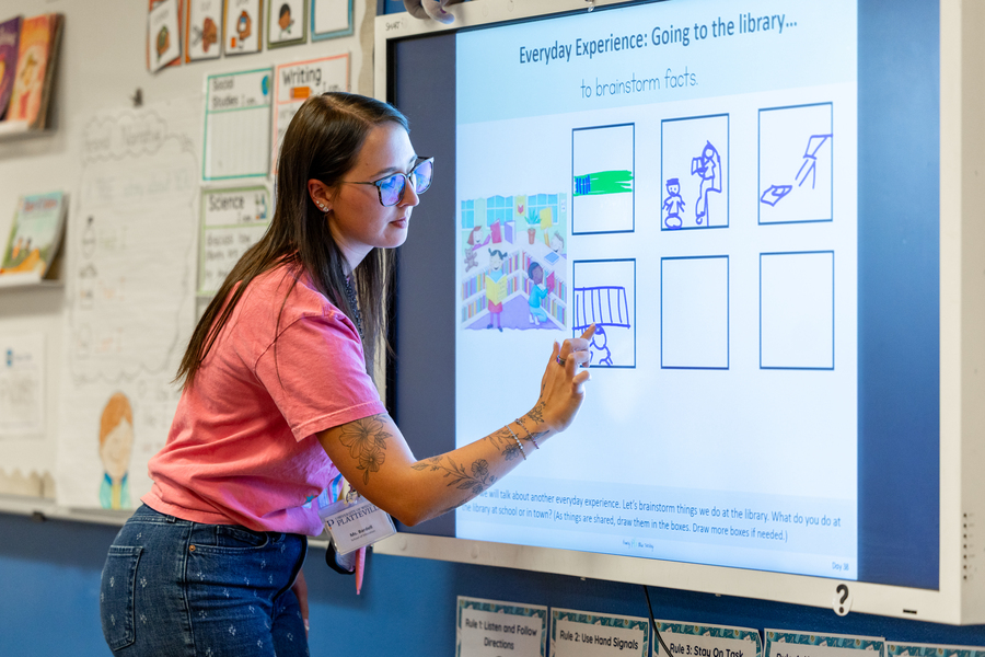 Educator interacting with a smartboard displaying a lesson about going to the library, highlighting activities to brainstorm facts.