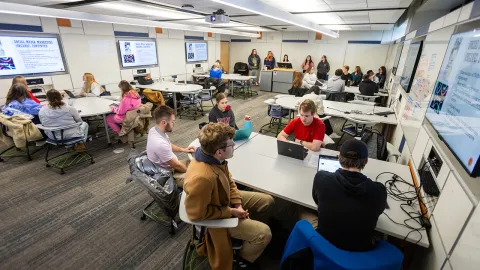 Students sitting in classroom with tables and screens on the wall.
