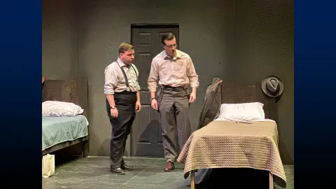 Two actors on stage next to a bed in the performance “The Dumb Waiter” by Harold Pinter.