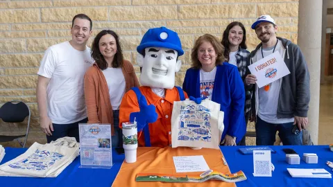 Group of individuals standing in front of a table with a mascot, smiling and holding signs for Pioneers Day of Giving