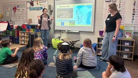 Two student teachers standing in a classroom in front of children seated on the floor