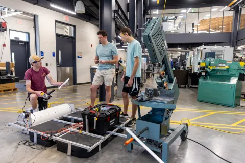 Students collaborating on a project in a workshop, discussing plans while working with equipment and tools on a workstation.