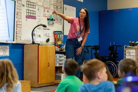 Teacher instructing students in a classroom, with a focus on an interactive board and educational materials in the background