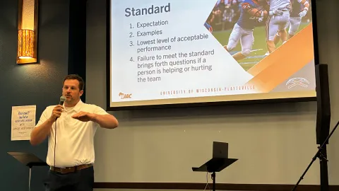 UW-Platteville Head Football Coach Ryan Munz speaks into a microphone in front of a screen in a banquet room as he discusses keys to leadership