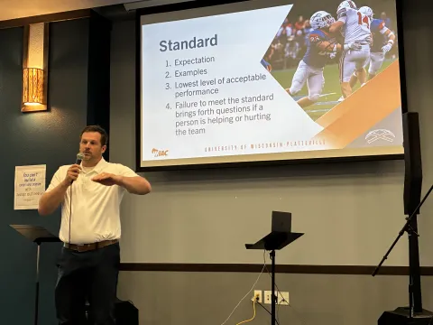 UW-Platteville Head Football Coach Ryan Munz speaks into a microphone in front of a screen in a banquet room as he discusses keys to leadership