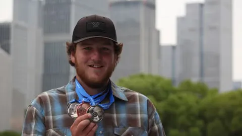UW-Platteville junior Jakob Scheckel wears a plaid shirt and cap as he holds up his medals standing in front of the downtown Nanjing, China, skyline.