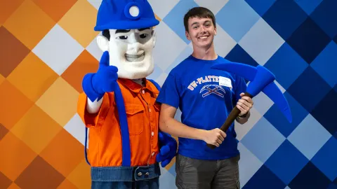 Person posing with Pioneer Pete, holding a blue pickaxe against a colorful geometric backdrop.