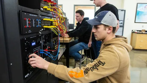 Students engaging in a hands-on electrical engineering lab, adjusting equipment with wires and control panels. 