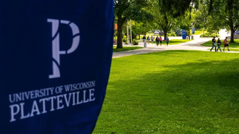 The image features a logo of the University of Wisconsin-Platteville prominently displayed in the foreground. In the background, a group of students walk on a campus sidewalk.