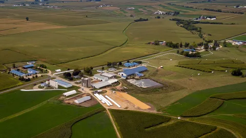 Aerial view of a rural farm landscape featuring multiple buildings, silos, fields filled with green crops.