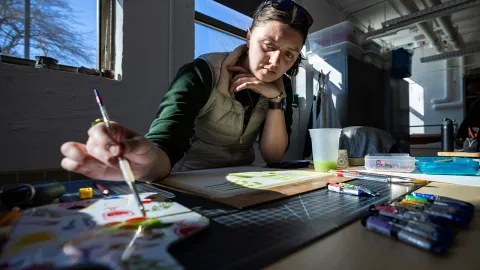 A student painting on a surface in a bright studio, with art supplies and tools visible on the table.