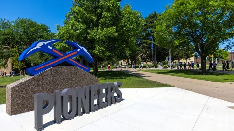 University of Wisconsin-Platteville signage displaying "Pioneers" with the crossed pickaxe logo, situated on campus with people walking along a path and trees in the background.