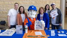 Group of individuals standing in front of a table with a mascot, smiling and holding signs for Pioneers Day of Giving