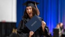 Smiling female student with dark hair wearing black graduation cap and gown, holding a diploma