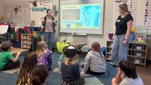 Two student teachers standing in a classroom in front of children seated on the floor