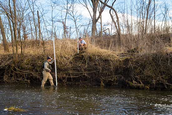 UW-Platteville student stands in river, measuring bank erosion while second student looks on.