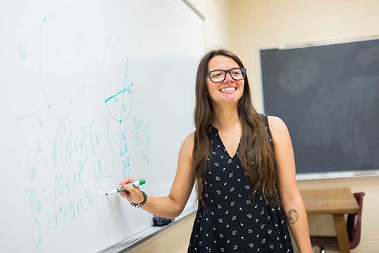 Smiling UW-Platteville student teacher stands at marker board writing equations.