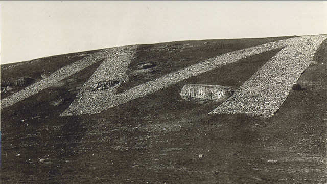 Landscape featuring a hillside displaying a large letter "M" made from white material against the sky.