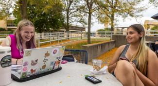 A UW-Platteville student sits with a laptop and notebook, laughing with a fellow student.
