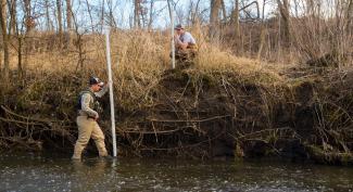 UW-Platteville student stands in river, measuring bank erosion while second student looks on.