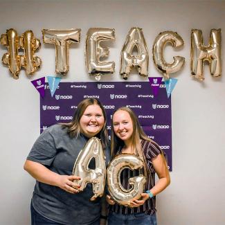 Two smiling students holding balloons of the letters A and G with #Teach above them