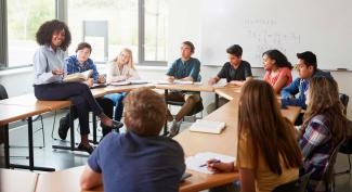 Smiling teacher sitting casually in front of a circle of young adults with a math equation on a whiteboard in the background.