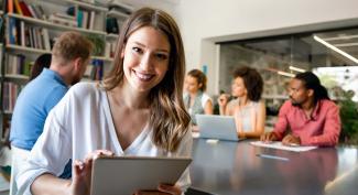 Smiling online student using a tablet at a meeting table with coworkers in the background.