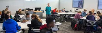 Students are seated at tables in a classroom setting, engaged in an interactive session with a faculty member, with interactive materials visible.
