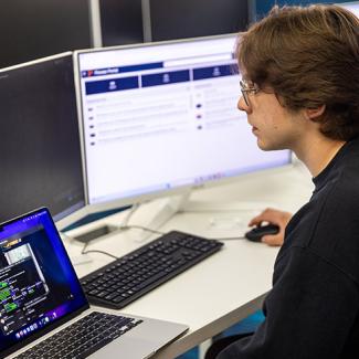 A student engaging in coding on a laptop while referencing data displayed on external monitors in a collaborative workspace.