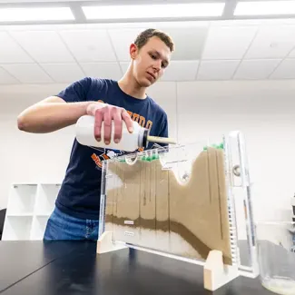 Student conducting a soil erosion experiment by pouring liquid into a transparent model with sand layers in a laboratory setting