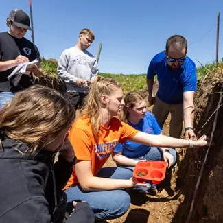Students observing soil analysis techniques in an outdoor classroom setting, with one holding a container of soil while another measures soil depth.