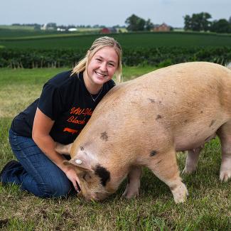 Young woman interacting with a pig while kneeling in a grassy field with a farm landscape in the background.